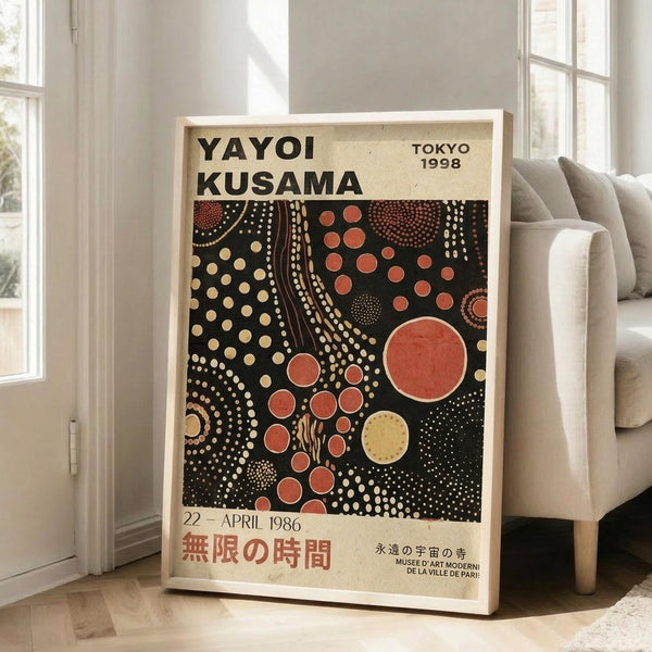 May include: Framed exhibition poster featuring Yayoi Kusama's signature polka dot artwork in black, red, orange and cream colors, displayed on wooden floor next to beige armchair in bright room with white walls and windows. The vintage-style poster shows circular dot patterns and museum exhibition text in both English and Japanese characters.