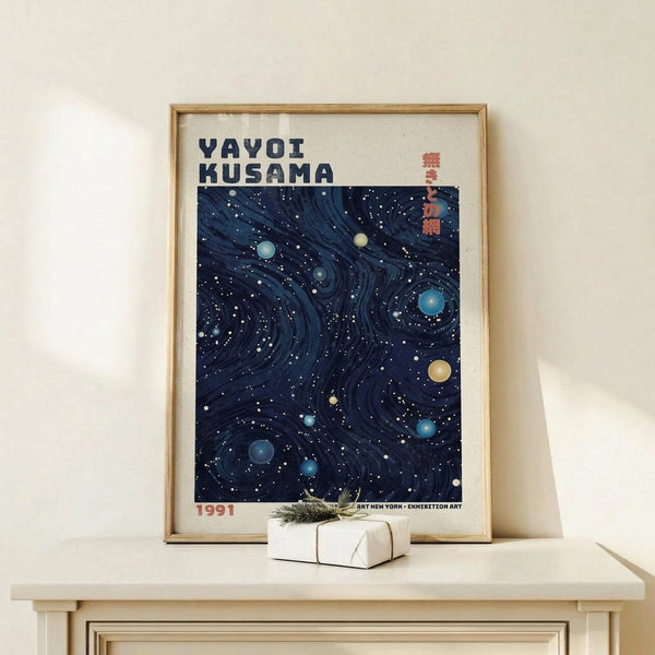 May include: Framed poster displaying swirling dark blue cosmic pattern with scattered colorful dots resembling planets and stars. Bold black text reads "YAYOI KUSAMA" at top with red Japanese characters and "1991" at bottom. Light wood frame sits on pale wooden surface next to small white gift box tied with twine and evergreen sprig against cream wall.