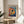 May include: Framed art print featuring a vibrant, colorful fish with polka dots in Kusama's distinctive style, displayed on a light wood round side table next to a beige upholstered chair. The artwork shows a stylized fish with a prominent dark eye surrounded by multicolored dots in blues, reds, yellows, and oranges against a dotted background. Japanese text appears below the image. Natural lighting creates soft shadows in the minimalist room setting with white walls and a plant visible in the