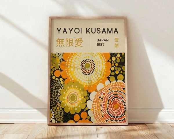 May include: Framed art poster featuring Yayoi Kusama's signature polka dot style with large circular flower-like patterns in orange, yellow, and green tones against a dark background. The cream-colored poster displays "YAYOI KUSAMA" at the top with Japanese characters and "JAPAN 1987" below. The wooden frame leans against a white wall on light hardwood flooring with soft natural lighting creating diagonal shadows across the scene.