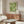 May include: Green and white dotted pattern artwork with "YAYOI KUSAMA TOKYO" text displayed in thin wooden frame above mid-century walnut credenza. Modern living room setting features white curved chair, glass oval coffee table, brass floor lamp, and soft natural lighting creating shadows on neutral walls.