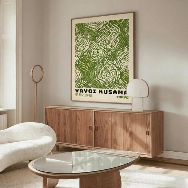 May include: Green and white dotted pattern artwork with "YAYOI KUSAMA TOKYO" text displayed in thin wooden frame above mid-century walnut credenza. Modern living room setting features white curved chair, glass oval coffee table, brass floor lamp, and soft natural lighting creating shadows on neutral walls.