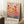 May include: Framed art poster leaning against white wall beside wooden side table with coffee cup. The artwork features colorful abstract botanical design with circular shapes resembling flowers or balloons in warm reds, oranges, and cool blues on cream background. Text reads "Yayoi Kusama" with Japanese characters above. Natural wood frame, positioned on white textured rug next to beige fabric in soft daylight.