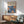May include: Framed poster with Yayoi Kusama text and Japanese characters at top, featuring blue and orange abstract floral or cellular patterns below. Displayed on white wall above wooden sideboard with small ceramic lamp. Mid-century modern green upholstered chair with wooden arms visible in foreground. Natural sunlight creates shadows across the minimalist room setting.