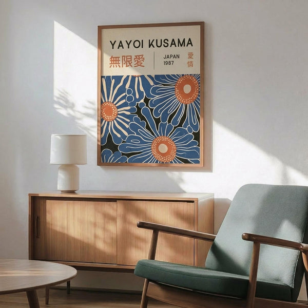 May include: Framed poster with Yayoi Kusama text and Japanese characters at top, featuring blue and orange abstract floral or cellular patterns below. Displayed on white wall above wooden sideboard with small ceramic lamp. Mid-century modern green upholstered chair with wooden arms visible in foreground. Natural sunlight creates shadows across the minimalist room setting.