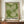 May include: Framed poster with green and cream dotted pattern resembling organic cellular forms or botanical motifs, displayed on wooden sideboard next to white ceramic vase and stacked books. The artwork features flowing, radiating dot patterns in various shades of green against cream background, with black text at bottom including Japanese characters. Warm interior setting with beige walls and crown molding.