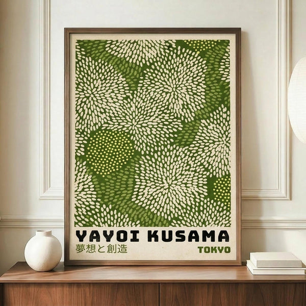 May include: Framed poster with green and cream dotted pattern resembling organic cellular forms or botanical motifs, displayed on wooden sideboard next to white ceramic vase and stacked books. The artwork features flowing, radiating dot patterns in various shades of green against cream background, with black text at bottom including Japanese characters. Warm interior setting with beige walls and crown molding.