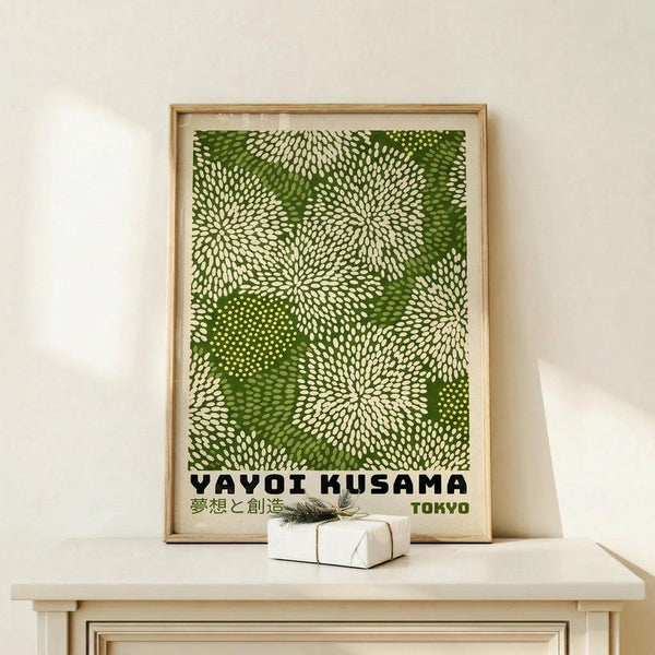 May include: Framed green and white dotted pattern artwork with radiating circular designs and Japanese text, displayed on light wood surface next to small wrapped gift with twine and evergreen sprig against cream wall