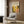 May include: Framed art print featuring colorful circular dot patterns in yellow, orange, and black tones displayed on wood paneled wall above beige upholstered chair with geometric pillow. Modern living room setting with gold floor lamp, dark side table with open book, and natural lighting from window with curtains.