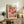 May include: Framed colorful abstract art poster displayed on rustic wooden credenza in modern living room setting. Artwork features vibrant pink, coral, orange and teal organic shapes with dotted patterns. Natural wood frame complements warm interior with fern plant, ceramic vase, stacked books, and neutral upholstered furniture visible.