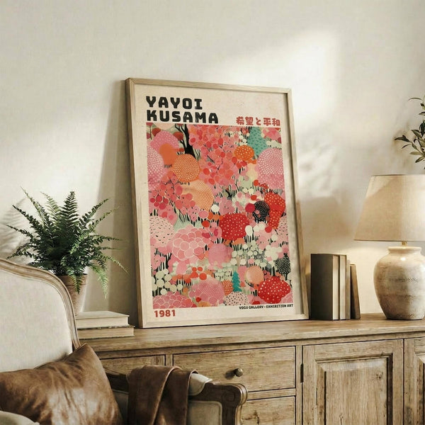 May include: Framed colorful abstract art poster displayed on rustic wooden credenza in modern living room setting. Artwork features vibrant pink, coral, orange and teal organic shapes with dotted patterns. Natural wood frame complements warm interior with fern plant, ceramic vase, stacked books, and neutral upholstered furniture visible.