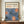 May include: Framed art print featuring blue stylized flowers with coral-orange centers and white dotted details on cream background, displayed on wooden sideboard next to white ceramic vase and stacked books against white paneled wall