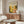 May include: Yayoi Kusama exhibition poster with colorful polka dot circles in yellow, orange, and green tones, displayed above a mid-century modern walnut credenza in a minimalist living room with white curved chair, glass oval coffee table, and soft natural lighting from window