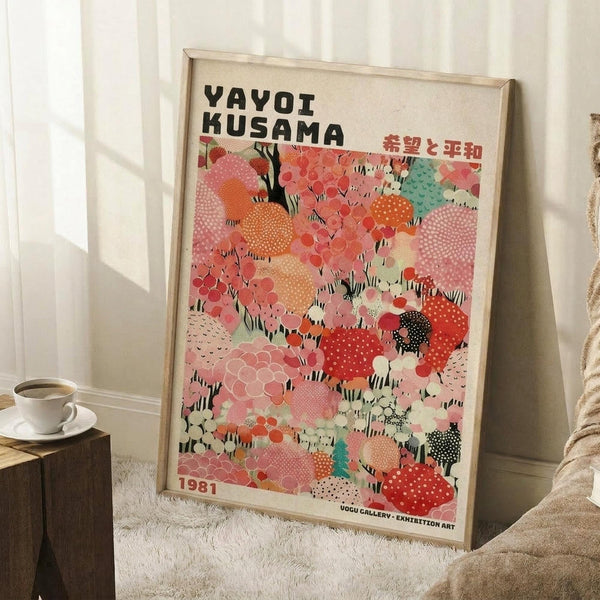 May include: Framed art poster leaning against white wall next to beige armchair and wooden side table with coffee cup. The colorful abstract artwork features organic blob shapes in coral, pink, orange, and teal with polka dot patterns. Black text at top reads "YAYOI KUSAMA" with Japanese characters. Bottom shows "1987" and gallery text. Light wooden frame, approximately 2-3 feet tall, displayed on white fluffy rug in bright room.
