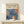 May include: Framed art print featuring abstract blue floral designs with orange dotted centers, displaying "YAYOI KUSAMA" text and Japanese characters at top with "JAPAN 1987". Natural wood frame leans against white wall on light wooden surface, with small white wrapped gift box decorated with greenery placed in front.