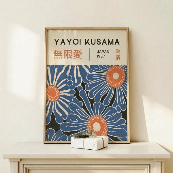 May include: Framed art print featuring abstract blue floral designs with orange dotted centers, displaying "YAYOI KUSAMA" text and Japanese characters at top with "JAPAN 1987". Natural wood frame leans against white wall on light wooden surface, with small white wrapped gift box decorated with greenery placed in front.