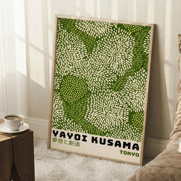 May include: Framed poster with green and white dotted pattern resembling organic cellular or floral forms, displayed in light wooden frame leaning against white wall. Scene shows cozy interior with coffee cup on wooden block, textured cream blanket, and natural window lighting creating soft shadows on wooden floor.