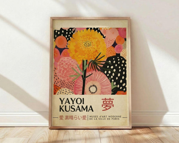 May include: Framed art print featuring vibrant abstract floral design with yellow dahlia, pink and coral organic shapes, black polka dot patterns, and radiating botanical forms. Poster displays "YAYOI KUSAMA" text and Japanese characters below colorful artwork. Natural wood frame sits on light wooden floor against white wall with soft window light creating diagonal shadows across the scene.