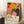 May include: Framed exhibition poster featuring vibrant botanical artwork with yellow sunflower, pink and coral abstract blooms, and black polka-dotted shapes against colorful background. Text shows artist name and museum details. Poster leans against white wall next to wooden side table with white coffee cup and saucer, beside beige knit throw blanket on wooden floor.