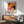 May include: Colorful Yayoi Kusama exhibition poster featuring vibrant organic shapes in orange, yellow, pink, and black with polka dot patterns, displayed in wooden frame above mid-century modern credenza. Scene shows mint green upholstered armchair with wooden arms, white ceramic table lamp, and sunny room with natural lighting casting shadows on white wall.
