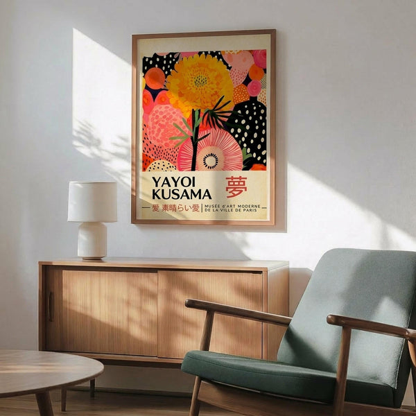 May include: Colorful Yayoi Kusama exhibition poster featuring vibrant organic shapes in orange, yellow, pink, and black with polka dot patterns, displayed in wooden frame above mid-century modern credenza. Scene shows mint green upholstered armchair with wooden arms, white ceramic table lamp, and sunny room with natural lighting casting shadows on white wall.