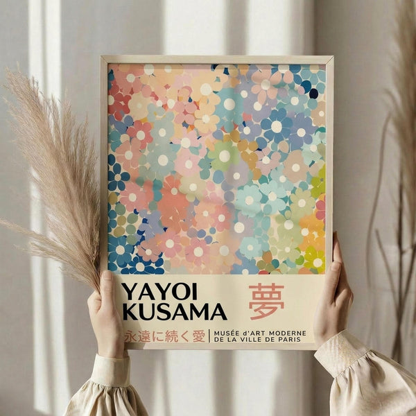 May include: Person holding framed art poster with colorful polka dot pattern in soft pastels - coral, blue, yellow, and white circles overlapping in dreamy watercolor style. Text reads "YAYOI KUSAMA" with Japanese characters and "MUSÉE d'ART MODERNE DE LA VILLE DE PARIS" below. Light wooden frame, neutral beige clothing, dried pampas grass visible, soft natural lighting against white curtained window background.