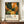 May include: Framed art poster featuring abstract botanical illustration with green foliage, orange and yellow organic shapes, white dots, and Japanese text reading "Yayoi Kusama" with "Musée d'Art Moderne de la Ville de Paris" below. Displayed on wooden sideboard next to white ceramic vase and books in modern room with white walls and paneled wainscoting.