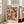 May include: Colorful framed poster featuring vibrant mushrooms and polka dot patterns in red, orange, pink, green, and yellow tones against black background. Japanese text at top. Light wood frame leaning against white wall next to beige upholstered chair in bright living room with large windows.