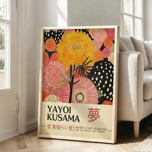 May include: A framed colorful botanical art print leaning against a white wall next to a beige upholstered sofa. The artwork features vibrant yellow flowers, pink blooms, and black polka dot patterns in a modern abstract style. Text at bottom reads "YAYOI KUSAMA" with Japanese characters and "MUSÉE D'ART MODERNE DE LA VILLE DE PARIS". Natural sunlight streams through nearby windows onto light wooden floors.