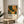 May include: Framed art print in light wood frame sitting on round wooden side table next to beige upholstered chair. Print features colorful abstract forest scene with green foliage, orange and yellow organic shapes, white dots scattered throughout, and text reading "YAYOI KUSAMA" with Japanese characters below. Soft natural lighting creates shadows across white wall. Plant leaves visible in corner.