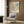 May include: Colorful abstract dotted artwork poster in light wood frame hanging on wood-paneled wall above beige upholstered armchair with geometric patterned pillow. Modern room setting with brass floor lamp, dark round side table with open book, and white curtains. Poster features multicolored circular pattern with Japanese text below.