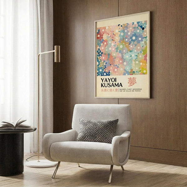 May include: Colorful abstract dotted artwork poster in light wood frame hanging on wood-paneled wall above beige upholstered armchair with geometric patterned pillow. Modern room setting with brass floor lamp, dark round side table with open book, and white curtains. Poster features multicolored circular pattern with Japanese text below.
