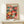 May include: A framed poster featuring colorful illustrated mushrooms in various sizes and patterns, displayed on a light wood console table with a small wrapped gift beside it. The artwork shows vibrant red, orange, pink, green, and yellow mushrooms with polka dots and organic shapes against a dark background, presented in a simple wooden frame against a cream wall.