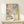 May include: Framed poster with colorful abstract polka dot pattern in pink, blue, and cream tones, featuring "YAYOI KUSAMA" text and Japanese characters below the artwork. Light wood frame leaning against white wall on pale wooden surface, with small white wrapped gift box tied with twine placed in front.