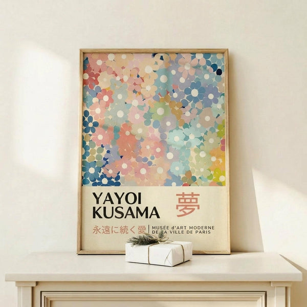 May include: Framed poster with colorful abstract polka dot pattern in pink, blue, and cream tones, featuring "YAYOI KUSAMA" text and Japanese characters below the artwork. Light wood frame leaning against white wall on pale wooden surface, with small white wrapped gift box tied with twine placed in front.
