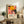 May include: Framed colorful poster with vibrant orange and pink floral patterns, black polka dot elements, and Japanese text, displayed on rustic wooden sideboard alongside fern plant, ceramic vase, and books in neutral-toned living room setting with beige chair visible.