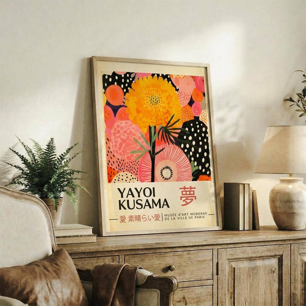May include: Framed colorful poster with vibrant orange and pink floral patterns, black polka dot elements, and Japanese text, displayed on rustic wooden sideboard alongside fern plant, ceramic vase, and books in neutral-toned living room setting with beige chair visible.