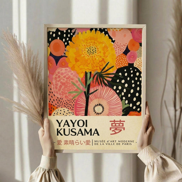 May include: Hands holding a framed poster featuring vibrant botanical artwork with a large yellow flower, coral and pink circular shapes, and black polka dot patterns against a cream background. The poster displays "YAYOI KUSAMA" text and Japanese characters. Setting shows neutral curtains and dried pampas grass in a minimalist room with natural lighting.