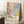 May include: Framed poster with colorful polka dot pattern in blues, pinks, corals, and greens displayed in bright room corner. White teacup on wooden block nearby, cream textured rug below, beige blanket draped beside frame. Text shows "YAYOI KUSAMA" with Japanese characters and "MUSEE d'ART MODERNE DE LA VILLE DE PARIS" in clean typography on cream background beneath dotted artwork.