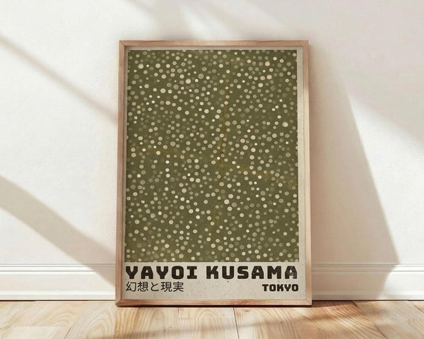 May include: Framed poster with olive green background covered in scattered cream and white polka dots of varying sizes, displayed against white wall on light wood flooring. Natural lighting creates soft shadows across the scene. Black text at bottom reads "YAYOI KUSAMA" with Japanese characters and "TOKYO" in smaller font. Light wooden frame surrounds the artwork.