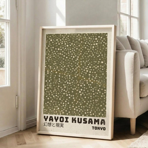 May include: Framed art print with olive green background covered in scattered cream and white polka dots of varying sizes, displayed in light wood frame leaning against white wall next to beige upholstered sofa. Print features black text reading "YAYOI KUSAMA" with Japanese characters and "TOKYO" below the dotted pattern. Bright natural light streams through tall windows in minimalist living room setting.