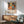 May include: Framed colorful abstract floral poster with "YAYOI KUSAMA JAPAN 1987" text hanging on white wall above wooden credenza with table lamp. Mid-century modern green upholstered chair with wooden arms in foreground. Artwork features vibrant flowers, polka dots, and organic shapes in orange, yellow, blue, and green tones. Natural sunlight creates shadows across the minimalist room setting.