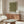May include: Modern living room with wood credenza displaying framed green poster featuring white polka dot pattern. Contemporary white sculptural chair and glass-top coffee table in foreground. Minimalist Scandinavian-style interior with neutral walls and natural lighting from window.
