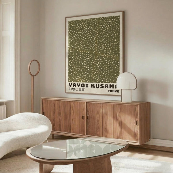 May include: Modern living room with wood credenza displaying framed green poster featuring white polka dot pattern. Contemporary white sculptural chair and glass-top coffee table in foreground. Minimalist Scandinavian-style interior with neutral walls and natural lighting from window.
