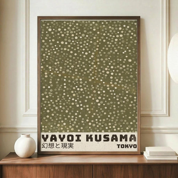May include: Framed poster with olive green background covered in scattered cream and white dots of varying sizes, displayed on wooden sideboard. Black text at bottom reads "YAYOI KUSAMA" with Japanese characters and "TOKYO". White ceramic vase and stacked books sit nearby against cream wainscoted wall.