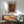 May include: Framed art poster with colorful abstract dotted pattern in yellows, oranges, purples and blues, displayed above a wooden sideboard with white ceramic lamp. Mid-century modern living room setting features mint green upholstered armchair with wooden frame, small round coffee table, and natural sunlight casting shadows on white wall.