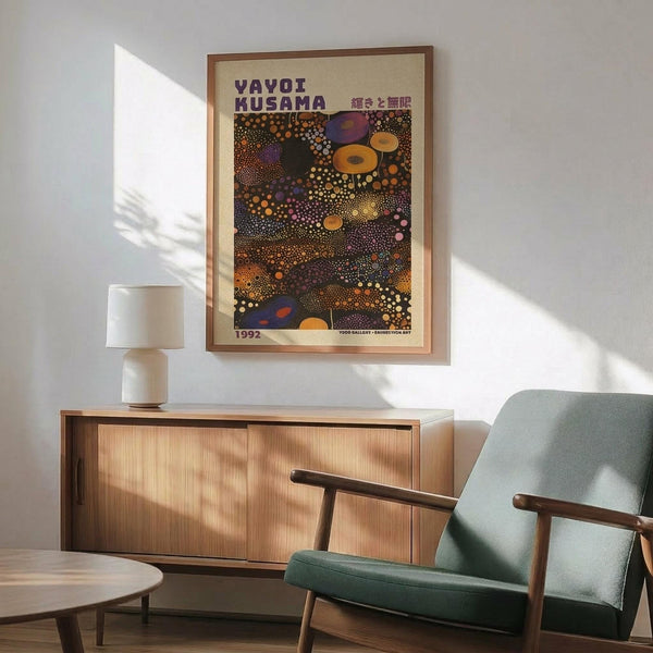 May include: Framed art poster with colorful abstract dotted pattern in yellows, oranges, purples and blues, displayed above a wooden sideboard with white ceramic lamp. Mid-century modern living room setting features mint green upholstered armchair with wooden frame, small round coffee table, and natural sunlight casting shadows on white wall.