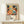May include: Framed art poster with colorful abstract floral design featuring large orange and yellow blooms, polka-dotted patterns in blues and greens, displayed on light wood console table next to small wrapped gift with twine and greenery sprig against cream wall