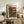 May include: Framed art print featuring colorful dotted abstract patterns in oranges, purples, and earth tones, displayed on rustic wooden sideboard alongside potted fern, ceramic vase, and stacked books in neutral-toned living room setting with cream chair and warm lighting.