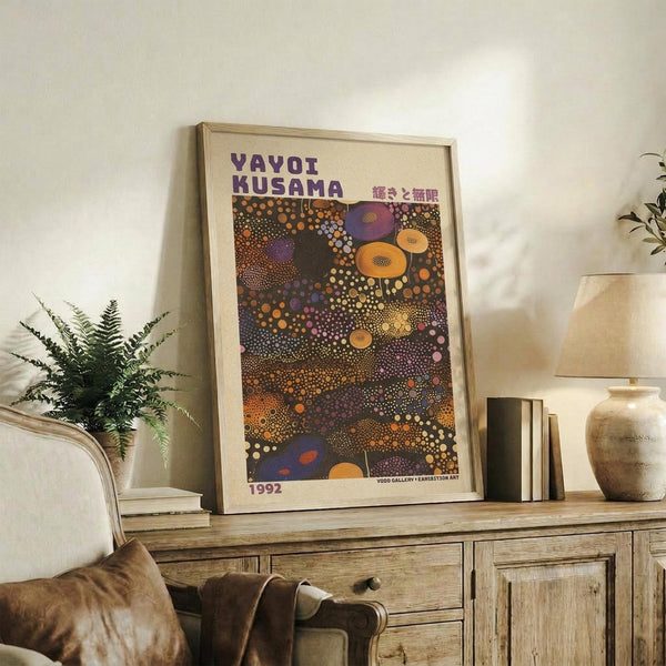 May include: Framed art print featuring colorful dotted abstract patterns in oranges, purples, and earth tones, displayed on rustic wooden sideboard alongside potted fern, ceramic vase, and stacked books in neutral-toned living room setting with cream chair and warm lighting.